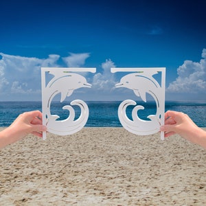 Peut inclure: Deux supports d'étagère en bois blanc en forme de dauphins sautant par-dessus des vagues. Les supports sont sur fond de ciel bleu et d'océan.