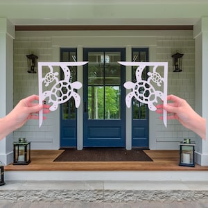 Könnte beinhalten: Weiße Metallhalterungen mit einem Meeresschildkröten-Design. Die Halterungen sind an der Seite einer blauen Tür mit Glasscheiben befestigt. Die Tür ist Teil einer weißen Veranda mit Holzboden.