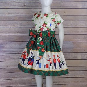 May include: A white dress with a green and white skirt. The skirt has a pattern of a nutcracker and other Christmas characters. The bodice of the dress has a pattern of poinsettias and holly.
