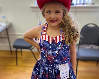 May include: A young girl in a red cowboy hat and red boots, wearing a blue dress with a white, red, and blue firework pattern. The dress has a white and red striped halter top and a tag that reads "Junior Miss Independence 22".