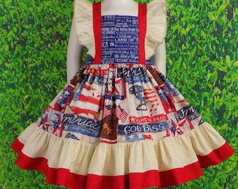 May include: A red, white, and blue dress with a ruffled skirt and a patriotic print. The dress features text such as "Free", "Born in the USA", "Home of the Brave", "Land of the Free", "God Bless", and "America".