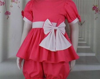 以下が含まれることがあります： ピンク色の子供用衣装。ワンピースとブルマのセット。ワンピースには白いリボンと、お揃いのリボン付きヘアバンドが付いています。衣装は白いトリムと白いベルトが特徴です。ブルマはフリル裾です。
