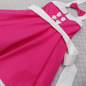 May include: A bright pink dress with white trim and accents. The dress features a white collar, straps, and bow details. A matching pink bow is also included. The dress is made of a smooth fabric.