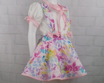 May include: A white blouse with puffed sleeves and a pink bow tie, paired with a pink and white floral print skirt with a pink bow at the waist. The skirt has a pink trim and a white lace trim on the blouse.