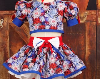 May include: A patriotic outfit featuring a crop top and skirt set with a matching bow. The fabric is patterned with red, white, and blue fireworks. The skirt has a white underskirt and a red waistband with a white bow. Blue bows adorn the shorts.