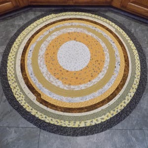 May include: A round braided rug with concentric circles in shades of yellow, brown, and gray. The center is white, surrounded by yellow, brown, and patterned bands. The outer edge is a dark gray.