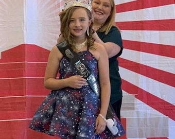 May include: A young girl wearing a blue and white dress with a star pattern and a crown on her head. She is holding a sash that says "America's Sweetheart". She is standing next to a woman wearing a green shirt. The background is a red, white, and blue striped backdrop with the word "Pageant" at the top.