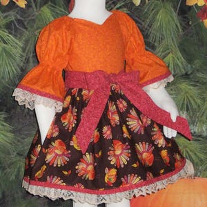 May include: A girl's dress with a bright orange top and a brown skirt with a turkey print. The skirt has a pink ribbon tied around the waist and a white lace trim.