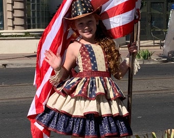 May include: A young person wearing a red, white, and blue dress with a ruffled skirt and a cowboy hat. They are holding a large American flag and wearing cowboy boots with a red, white, and blue pattern.