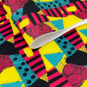 Pu&ograve; includere: Un tessuto colorato con un design a patchwork che presenta nero, giallo, rosa, blu e marrone. Il tessuto ha un motivo ripetitivo di forme geometriche e vortici.