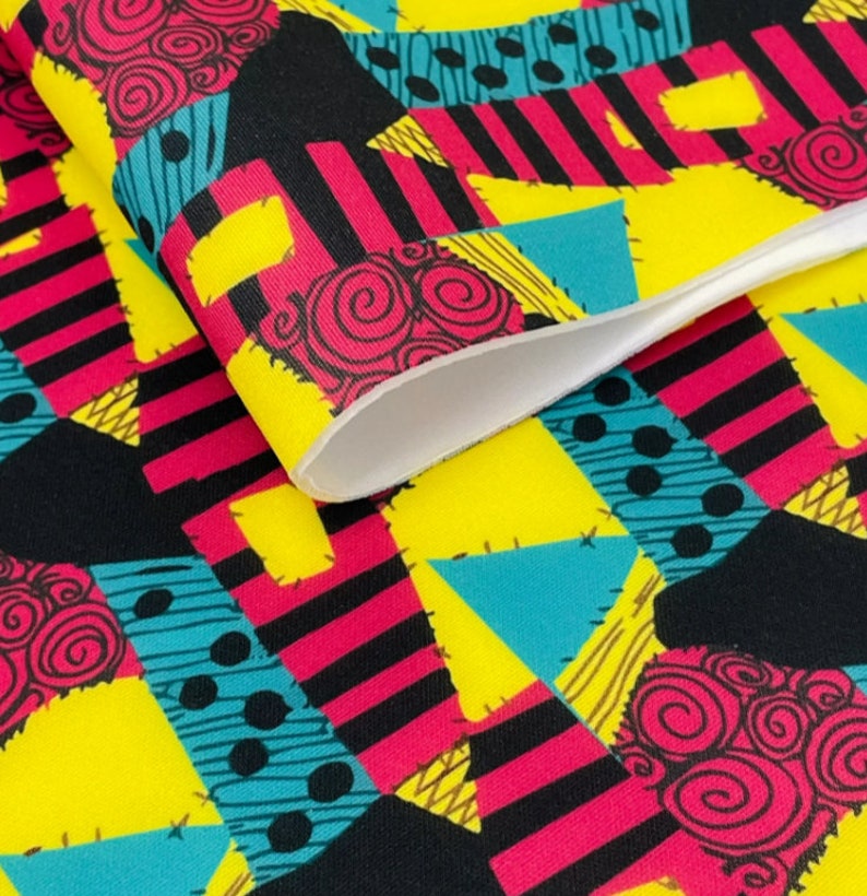 Pu&ograve; includere: Un tessuto colorato con un design a patchwork che presenta nero, giallo, rosa, blu e marrone. Il tessuto ha un motivo ripetitivo di forme geometriche e vortici.
