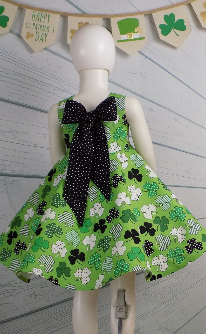 May include: A green dress with a shamrock print and a black polka dot bow in the back. The dress has a full skirt and is perfect for St. Patrick's Day.