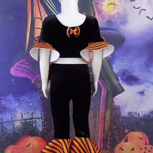 Pu&ograve; includere: Un costume di Halloween nero e arancione per un bambino. Il costume comprende un top corto in velluto nero con maniche a righe arancioni e nere, una gonna in velluto nero con bordatura a righe arancioni e nere e una fascia in velluto nero con un fiocco arancione.