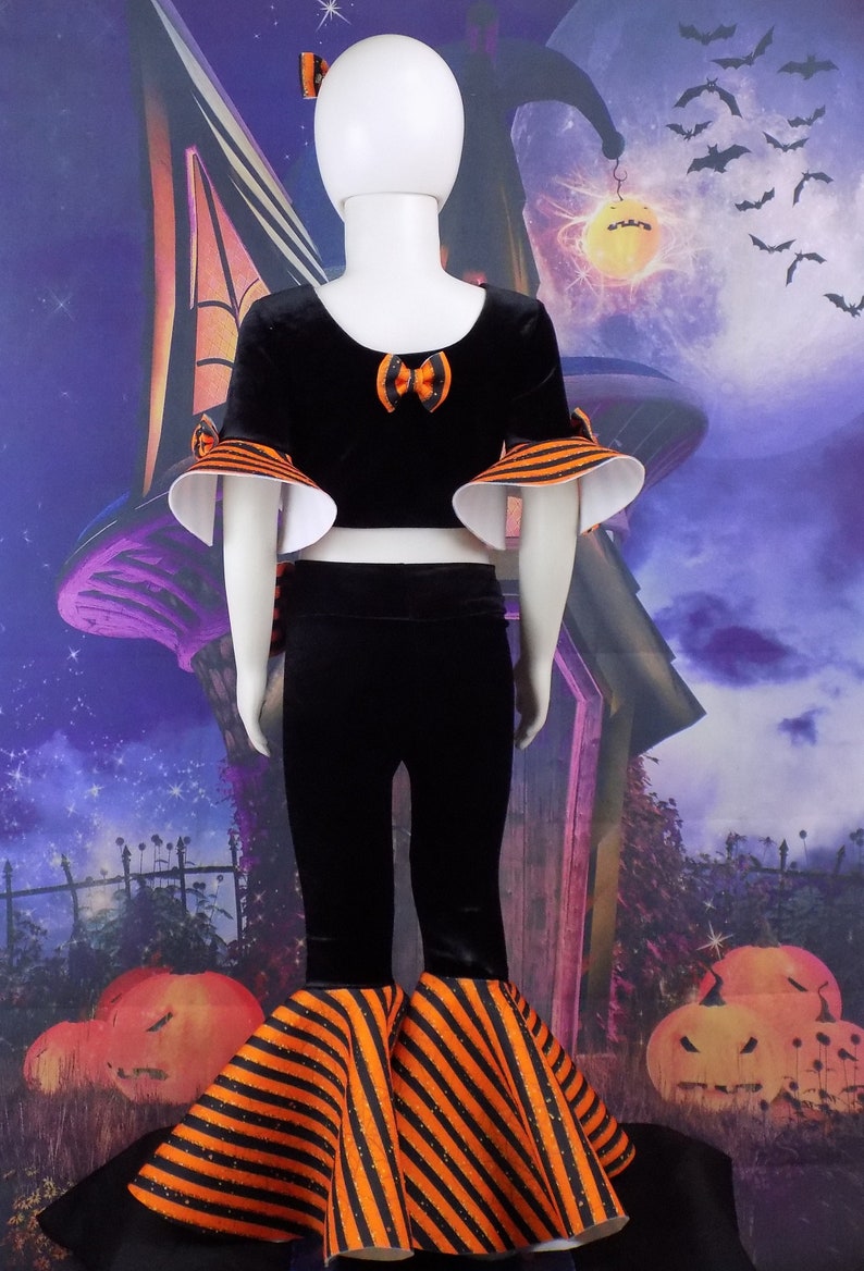 Pu&ograve; includere: Un costume di Halloween nero e arancione per un bambino. Il costume comprende un top corto in velluto nero con maniche a righe arancioni e nere, una gonna in velluto nero con bordatura a righe arancioni e nere e una fascia in velluto nero con un fiocco arancione.