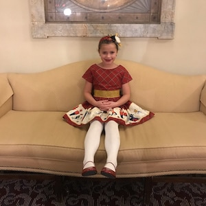 May include: A young girl wearing a red and white plaid dress with a gold belt and a white skirt with a colorful print sits on a beige couch. She is wearing white tights and red shoes.