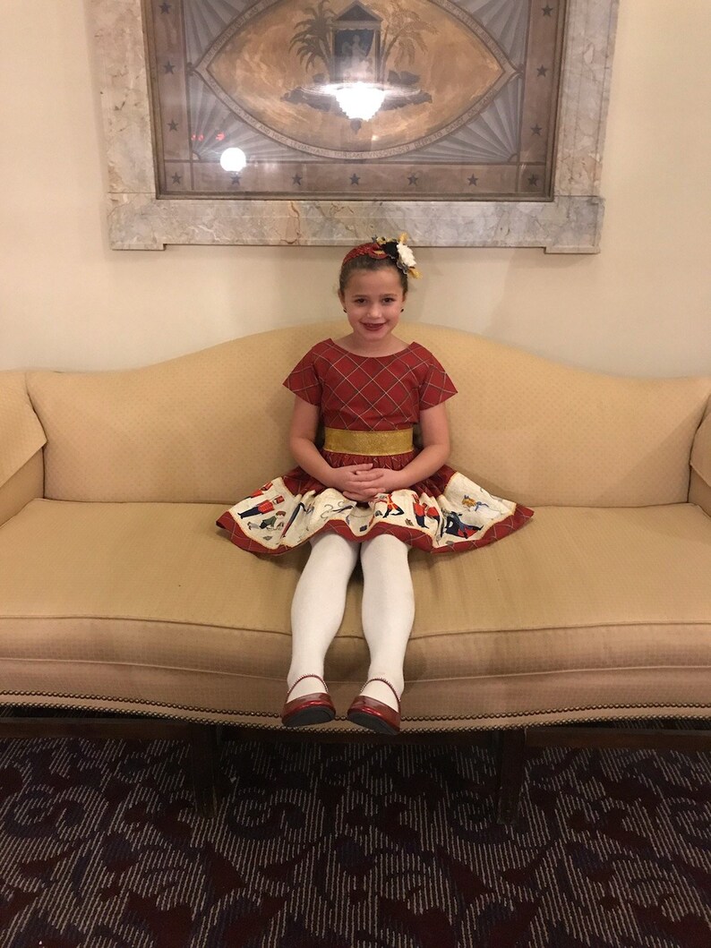 May include: A young girl wearing a red and white plaid dress with a gold belt and a white skirt with a colorful print sits on a beige couch. She is wearing white tights and red shoes.