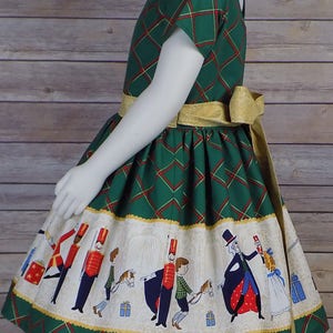 May include: A green plaid dress with a gold belt and a white skirt featuring a whimsical illustration of toy soldiers and children playing with toys. The dress has a festive Christmas theme.