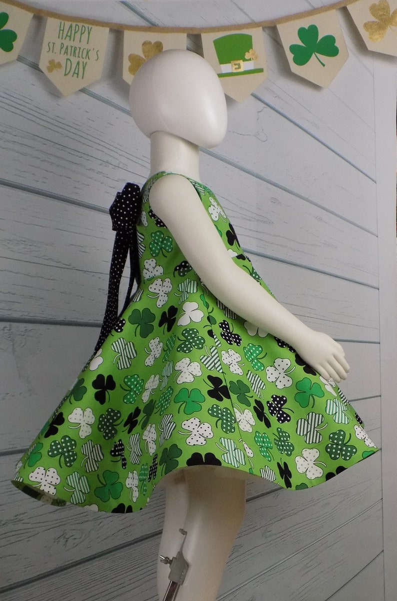 May include: A green sleeveless dress with a black and white polka dot bow in the back. The dress is covered in a pattern of green shamrocks and white polka dots on a green background. The dress is on a white mannequin.