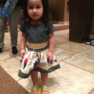 May include: A young girl wearing a green and white plaid dress with a gold belt and yellow shoes. The dress has a white skirt with a pattern of people and animals. The girl is standing on a tiled floor.