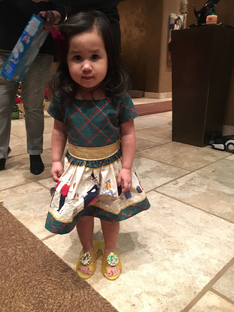 May include: A young girl wearing a green and white plaid dress with a gold belt and yellow shoes. The dress has a white skirt with a pattern of people and animals. The girl is standing on a tiled floor.
