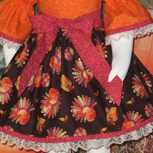 May include: A brown and orange dress with a turkey print and a pink bow. The dress has a white lace trim.