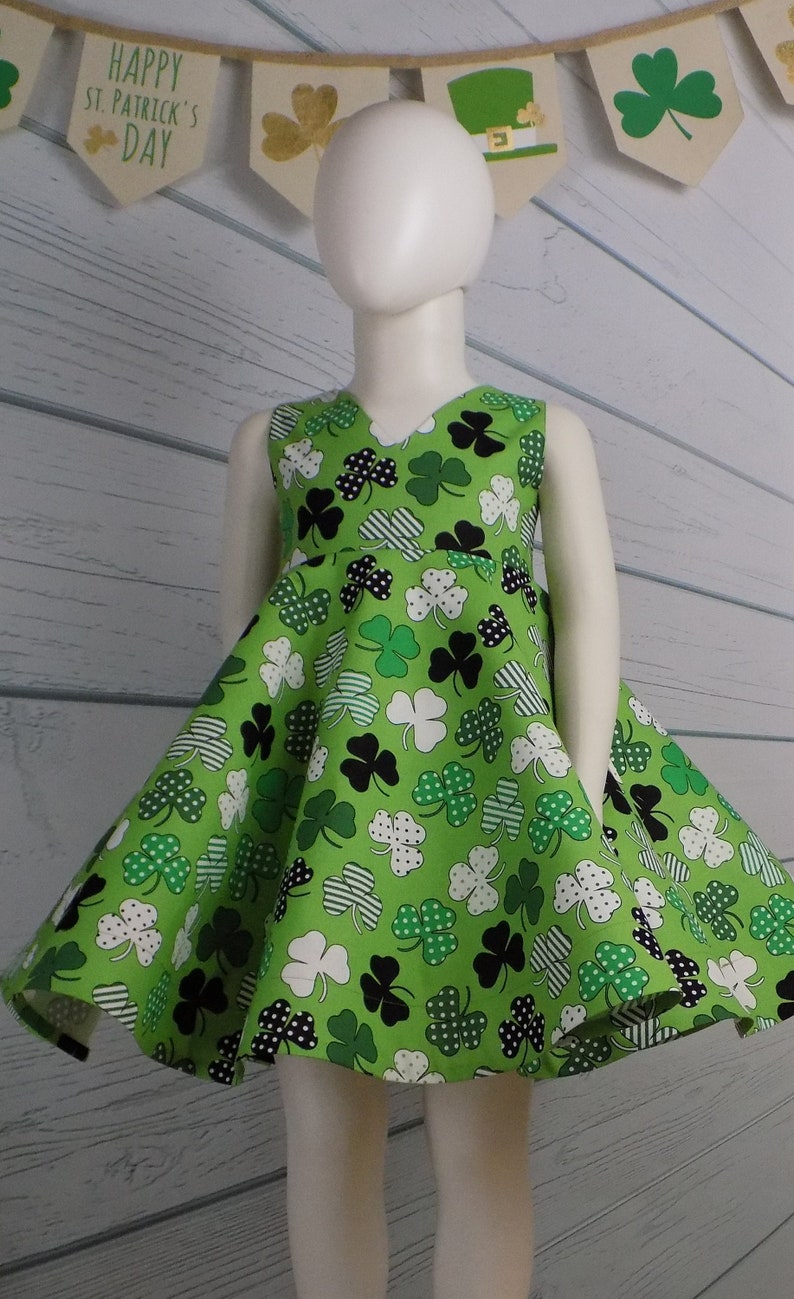 May include: A green sleeveless dress with a shamrock print. The dress has a V-neckline and a flared skirt. The dress is on a white mannequin.