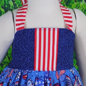 May include: A blue and white sleeveless dress with red, white, and blue striped straps. The dress has a blue bodice with a red, white, and blue striped panel and a blue skirt with a white and red print.