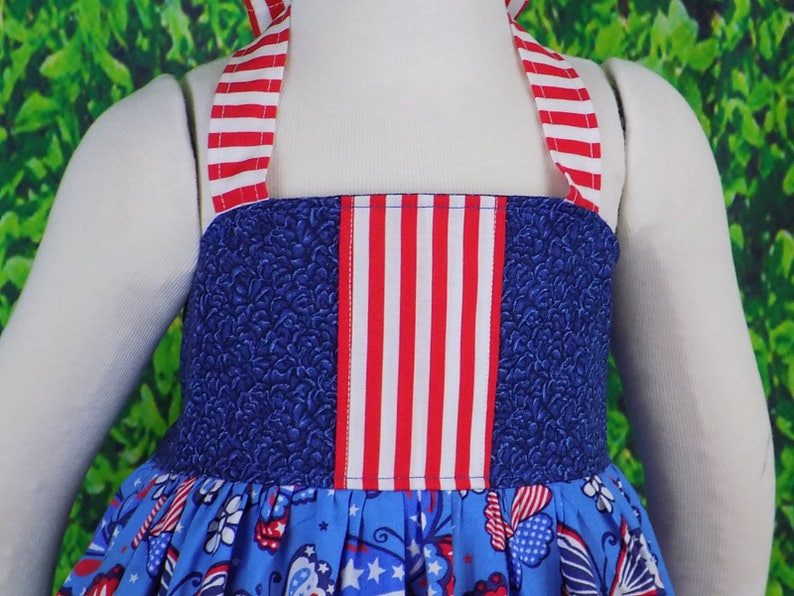 May include: A blue and white sleeveless dress with red, white, and blue striped straps. The dress has a blue bodice with a red, white, and blue striped panel and a blue skirt with a white and red print.