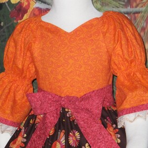 May include: A girl's dress with a brown and orange floral print. The dress has a pink ribbon tied around the waist and orange puffed sleeves with white lace trim.