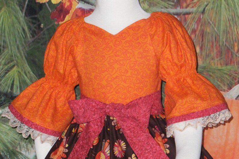May include: A girl's dress with a brown and orange floral print. The dress has a pink ribbon tied around the waist and orange puffed sleeves with white lace trim.