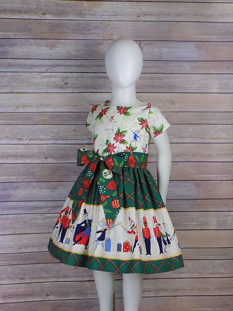 May include: A white dress with a green and white skirt. The skirt has a pattern of a nutcracker and other Christmas characters. The bodice of the dress has a pattern of poinsettias and holly.