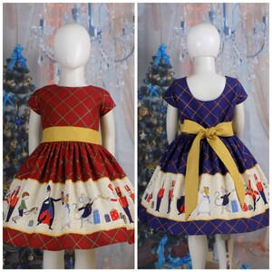 May include: Two girls wearing festive Christmas dresses with a Nutcracker theme. The dresses are red and blue with a white trim and feature a Nutcracker scene with toy soldiers, a mouse king, and a ballerina. The dresses have yellow sashes.