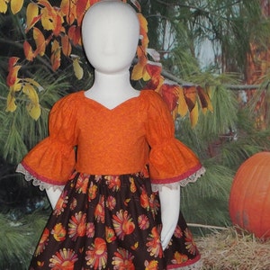 May include: A brown and orange dress with a turkey print and lace trim. The dress has a fitted bodice and a full skirt. The dress is on a white mannequin against a backdrop of fall leaves.