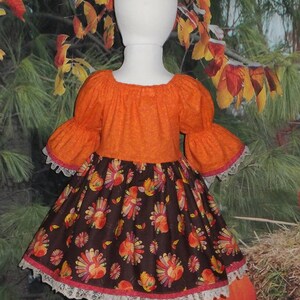 May include: A girl's orange and brown dress with a turkey print skirt and white lace trim. The dress has a gathered bodice and puffed sleeves.