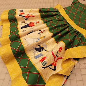 May include: A green and gold dress with a Nutcracker theme. The dress has a plaid pattern and a gold trim. The skirt features a Nutcracker scene with a soldier, a ballerina, and a mouse.