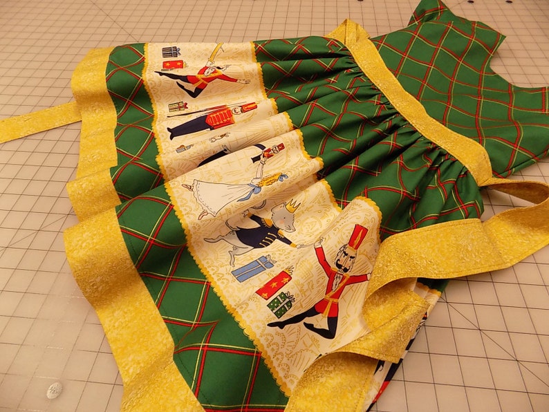 May include: A green and gold dress with a Nutcracker theme. The dress has a plaid pattern and a gold trim. The skirt features a Nutcracker scene with a soldier, a ballerina, and a mouse.