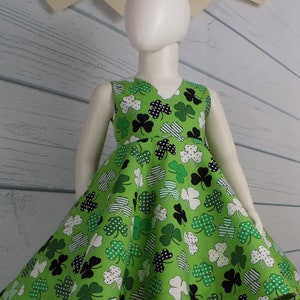 May include: A green sleeveless dress with a shamrock print. The dress has a V-neckline and a full skirt. The dress is on a white mannequin. The background is a white wood panel with a banner that says "Happy St. Patrick's Day".