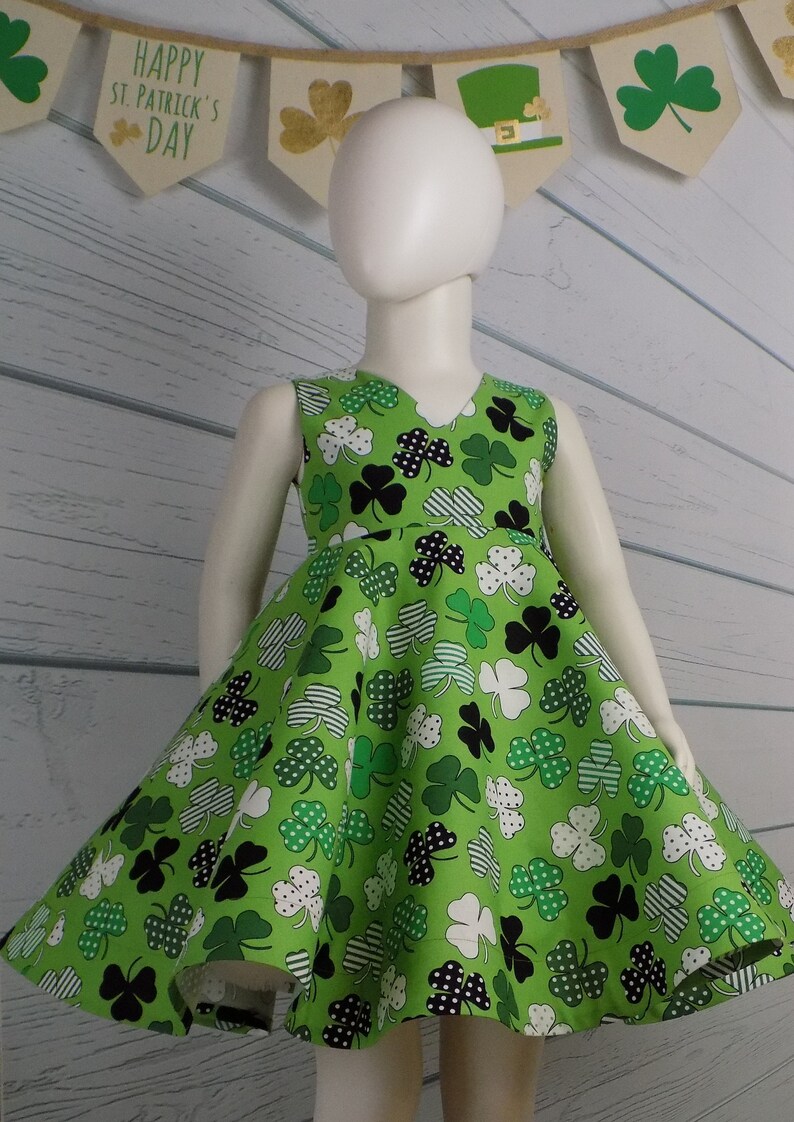 May include: A green sleeveless dress with a shamrock print. The dress has a V-neckline and a full skirt. The dress is on a white mannequin. The background is a white wood panel with a banner that says "Happy St. Patrick's Day".