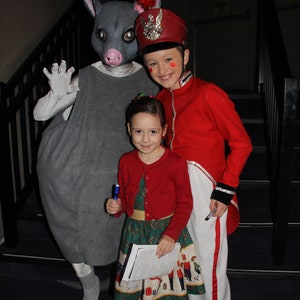 May include: A gray, furry, anthropomorphic animal wearing a gray dress stands with a girl wearing a red jacket and a white skirt. The girl is holding a blue pen. The girl is standing on a set of stairs. The animal has a red and white feathered hat on its head. The girl has a red and white feathered hat on her head. The girl is smiling. The animal is looking at the camera.