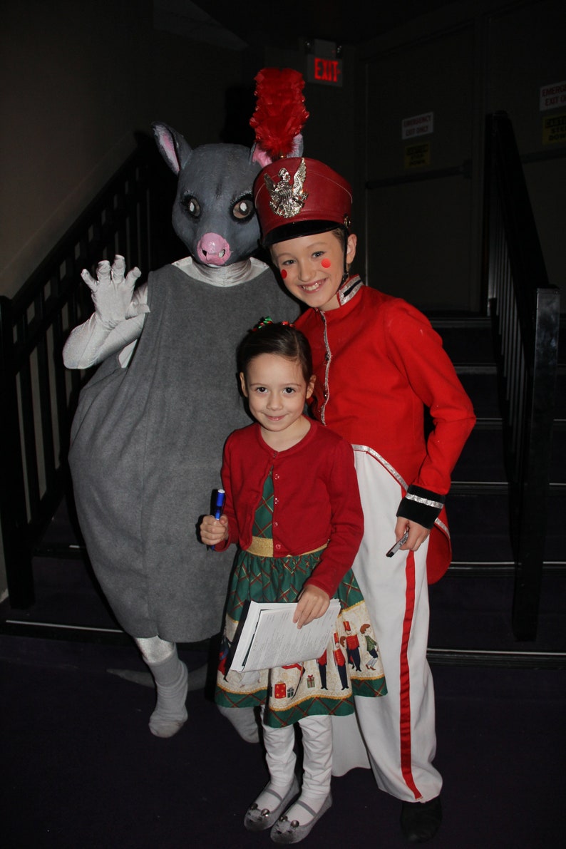 May include: A gray, furry, anthropomorphic animal wearing a gray dress stands with a girl wearing a red jacket and a white skirt. The girl is holding a blue pen. The girl is standing on a set of stairs. The animal has a red and white feathered hat on its head. The girl has a red and white feathered hat on her head. The girl is smiling. The animal is looking at the camera.