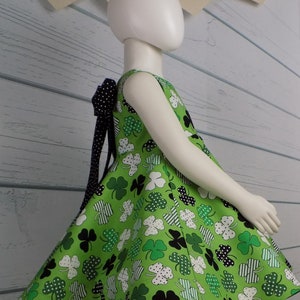 May include: A green sleeveless dress with a shamrock print and a black polka dot bow. The dress has a full skirt and is perfect for a St. Patrick's Day celebration. The banner above reads "Happy St. Patrick's Day".