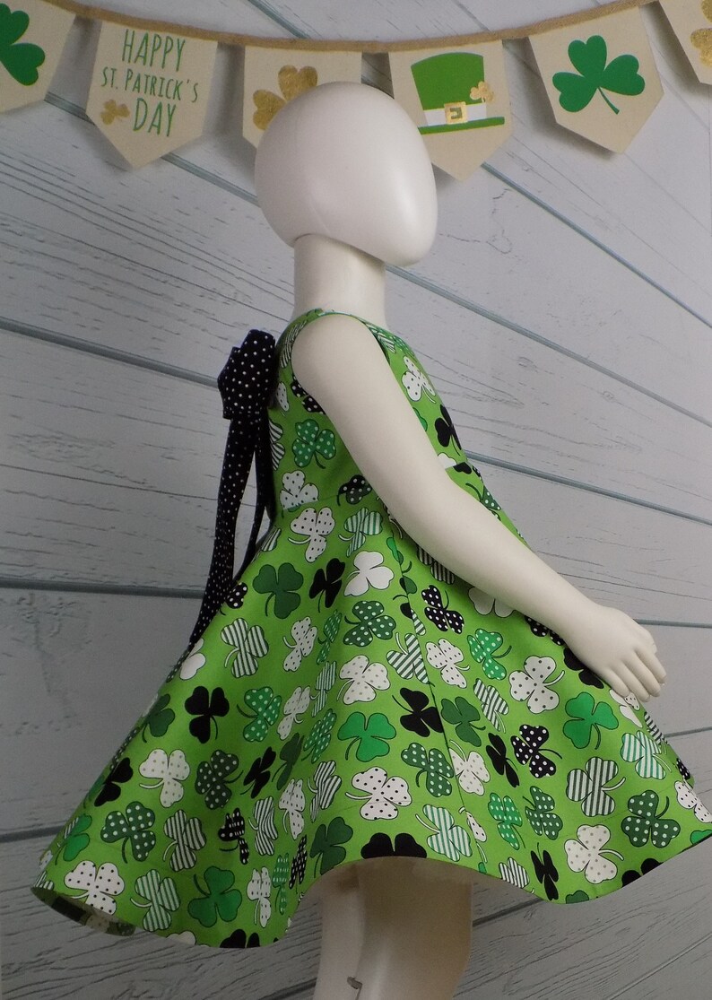 May include: A green sleeveless dress with a shamrock print and a black polka dot bow. The dress has a full skirt and is perfect for a St. Patrick's Day celebration. The banner above reads "Happy St. Patrick's Day".