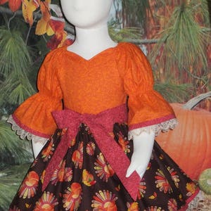 May include: A brown and orange dress with a turkey print and a pink ribbon. The dress has a lace trim and is worn by a mannequin.