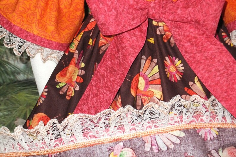 May include: A brown fabric with a pattern of colorful turkeys and flowers. The fabric has a pink ribbon tied around it and a white lace trim.
