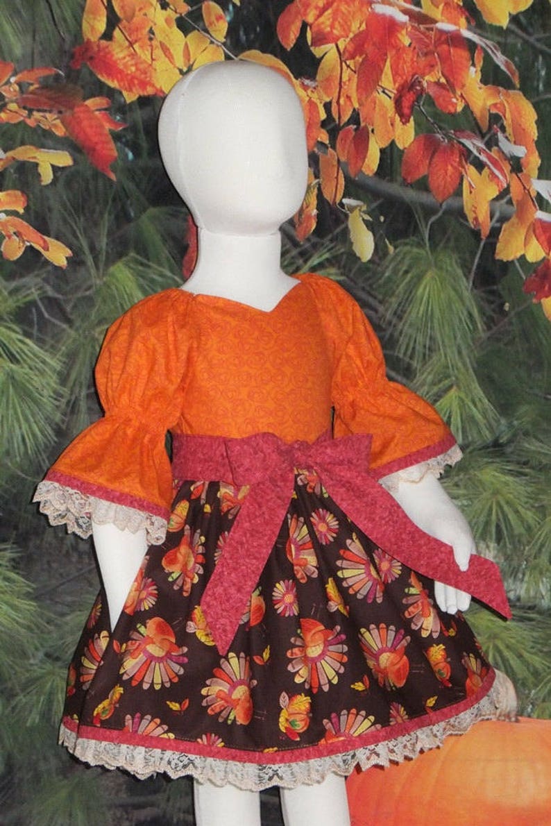 May include: A girl's dress with a bright orange top and a brown skirt with a turkey print. The skirt has a pink ribbon tied around the waist and a white lace trim.