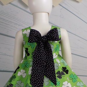 May include: A green dress with a shamrock print and a black bow in the back. The dress has a full skirt and is sleeveless. The dress is perfect for St. Patrick's Day.