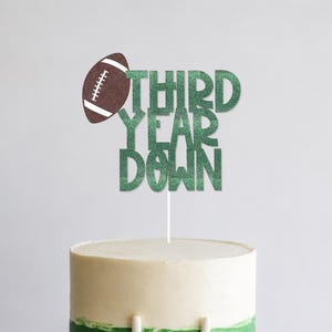 May include: A cake decorated for a football-themed celebration. The cake has a white and green color scheme, with a football and a goalpost made of icing. A cake topper reads "THIRD YEAR DOWN" in green glitter.