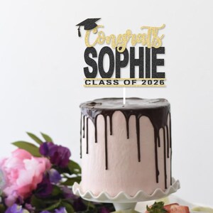 May include: A graduation cake with pink frosting and chocolate ganache drips. A cake topper reads "Congrats Sophie Class of 2026" in black and gold glitter. The cake sits on a white scalloped cake stand, with flowers and strawberries in the background.