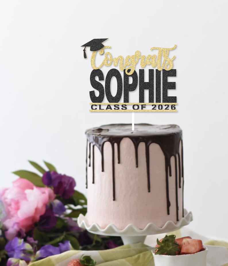 May include: A graduation cake with pink frosting and chocolate ganache drips. A cake topper reads "Congrats Sophie Class of 2026" in black and gold glitter. The cake sits on a white scalloped cake stand, with flowers and strawberries in the background.