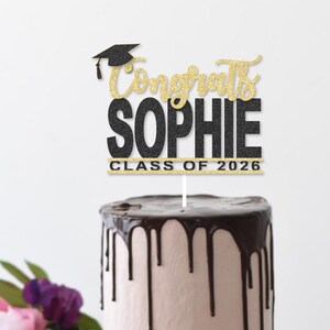 May include: A cake topper with the words "Congrats Sophie" in gold glitter script and black block letters, topped with a graduation cap. The text "Class of 2026" is also included. The topper is placed on a cake with chocolate frosting.